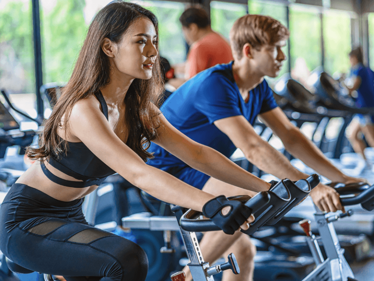 Une femme et un homme sont engagés dans un cours de RPM, concentrés sur leur entraînement cardio sur des vélos de spinning dans une salle de sport spacieuse et bien éclairée de l'Appart Fitness.