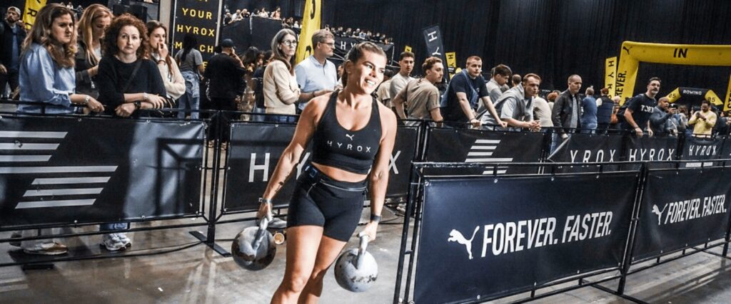 Participante Hyrox portant des kettlebells devant les supporters, lors d'une compétition grâce à un entraînement à L'Appart Fitness.